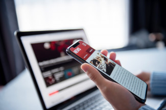 A hand holding a cell phone reading news headlines, with a laptop open in the background.