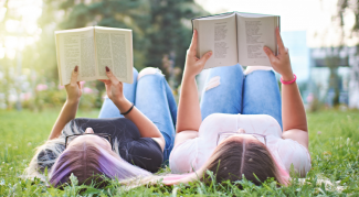 Two young women laying on their backs in the grass on a sunny day, holding books of poetry above their heads as they read.