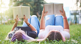 Two young women laying on their backs in the grass on a sunny day, holding books of poetry above their heads as they read. 