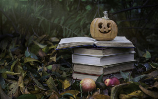 An autumnal outdoor scene of leaf-strewn grass with apples on the ground, a stack of books and a small jack-o-lantern on top of the books