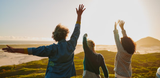 Three women seen from behind raise their arms joyfully toward the sun while standing on a grassy hill overlooking the ocean.